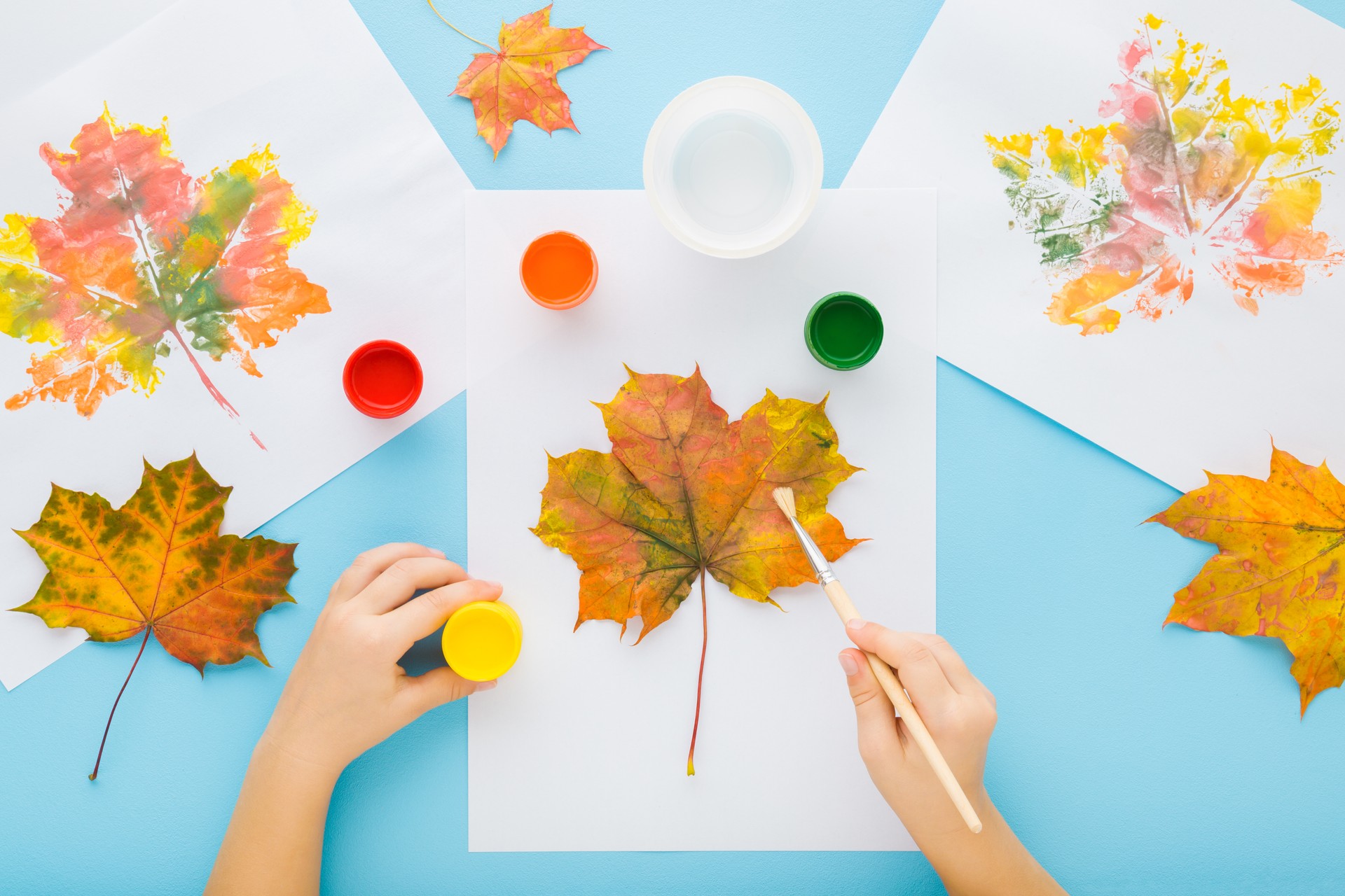 Little child hands painting and copying colorful leaves pattern with gouache color on white papers. Light blue table background. Pastel color. Closeup. Point of view shot. Top down view.