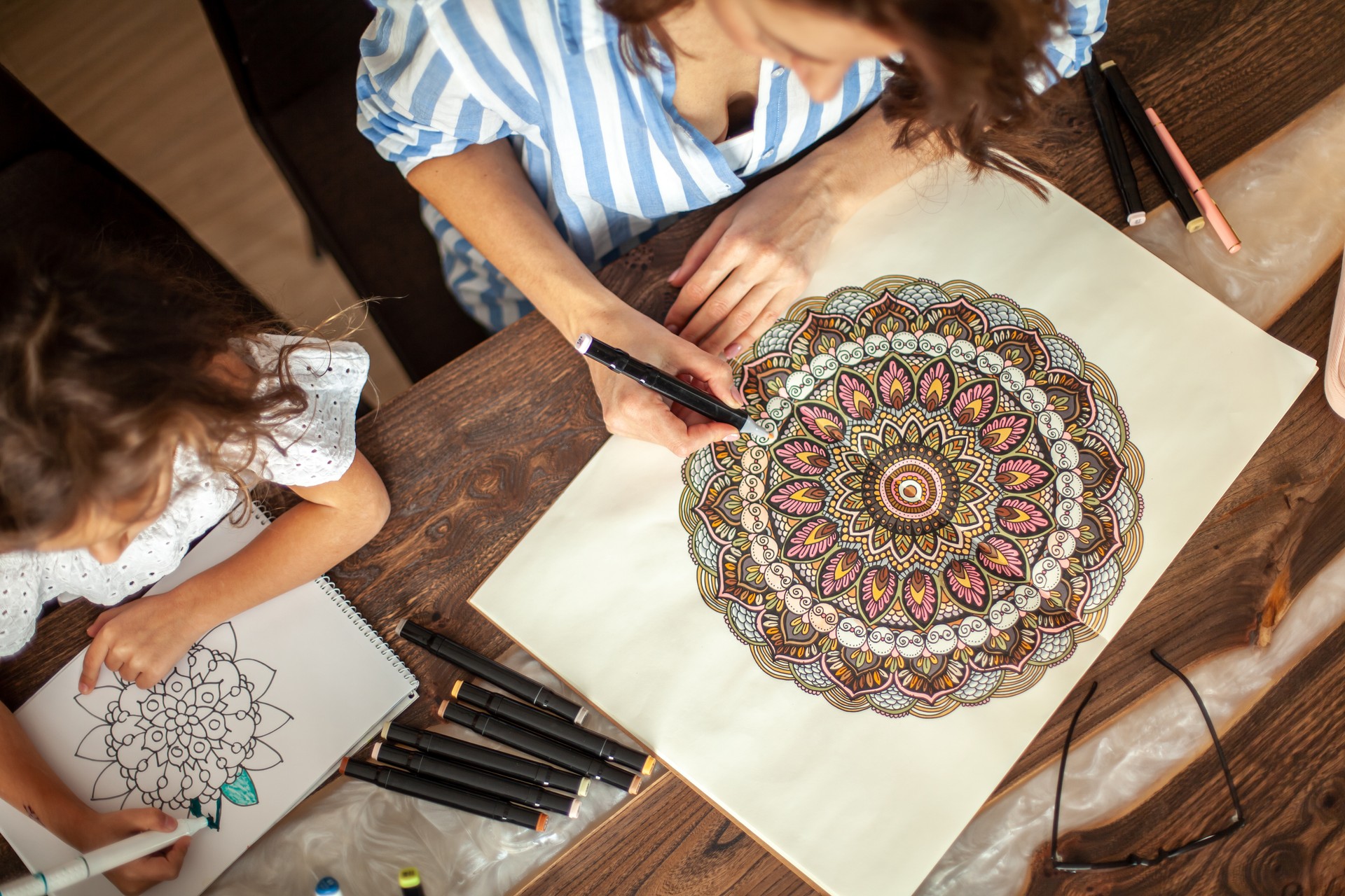 Young beautiful mother and cute daughter draw a mandala pattern together. Artistic markers and colored pencils